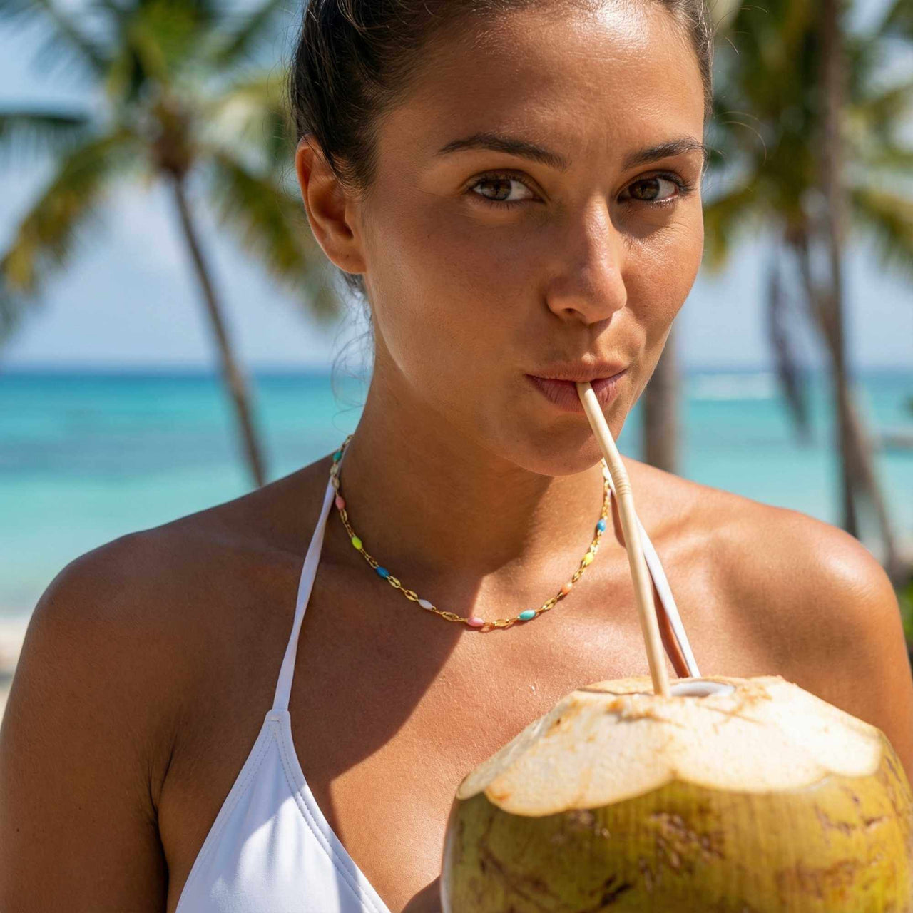 A woman enjoying a coconut drink while wearing a Rainbow Choker by Brother & Sisters on a beach.