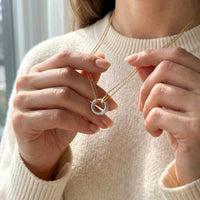Thumbnail for A woman showcases The Unity Bond necklace by Brother & Sisters, featuring a silver circle and gold bar pendant.