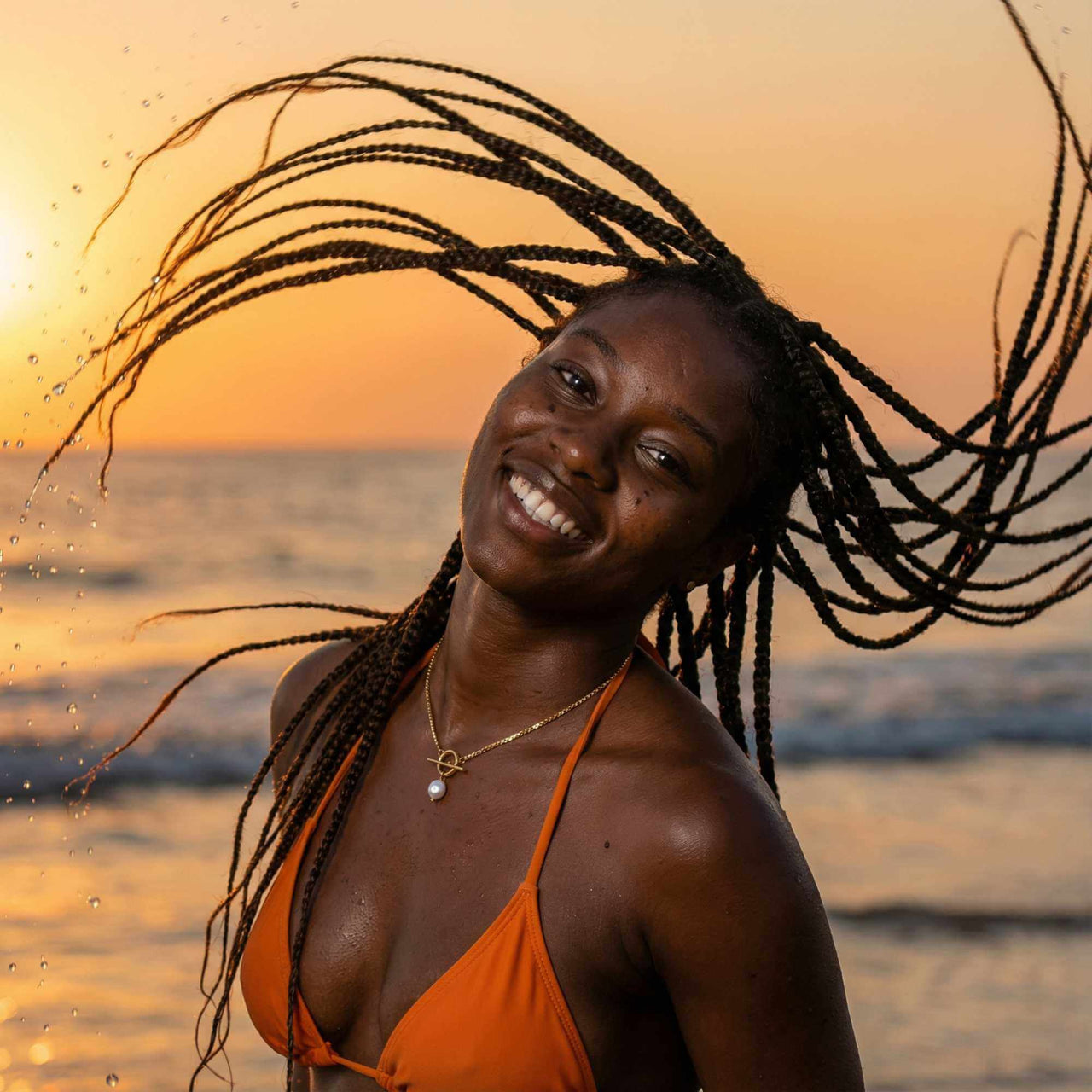 Happy woman with braided hair wearing La Isla Pearl Toggle Necklace, smiling at sunset by the sea.