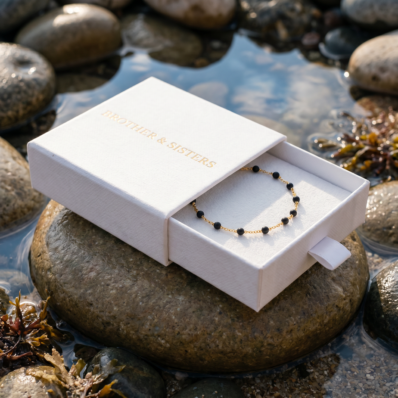 Gold & Onyx Bead Anklet in a luxurious Brother & Sisters box by the water, showcasing its elegance and sophistication.