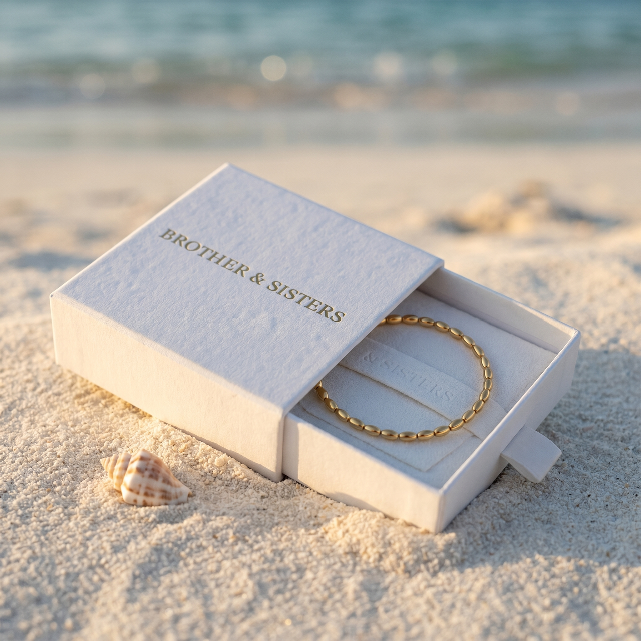 Oval Bead Anklet by Brother & Sisters in a box on the beach, showcasing gold beads and luxury packaging.