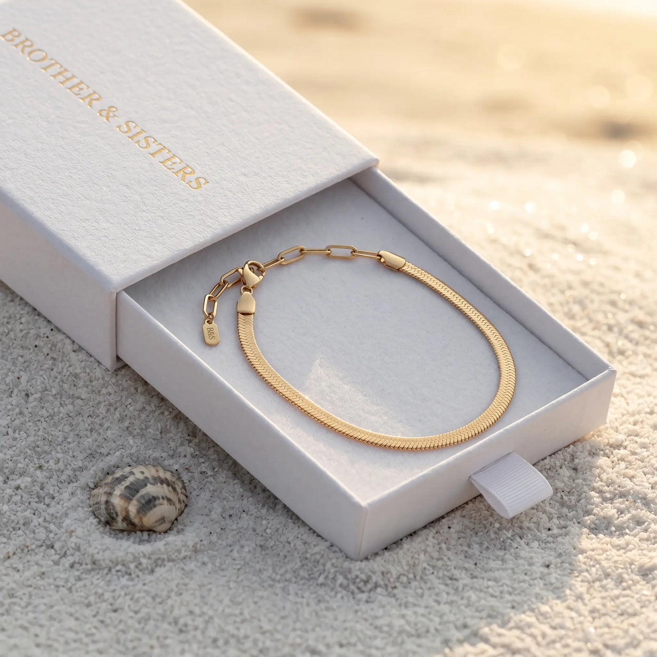 Layla gold anklet inside B&S signature white slide-drawer gift box on beach sand at golden hour with sea shell — flat herringbone snake chain visible as continuous oval, luxury anklet packaging