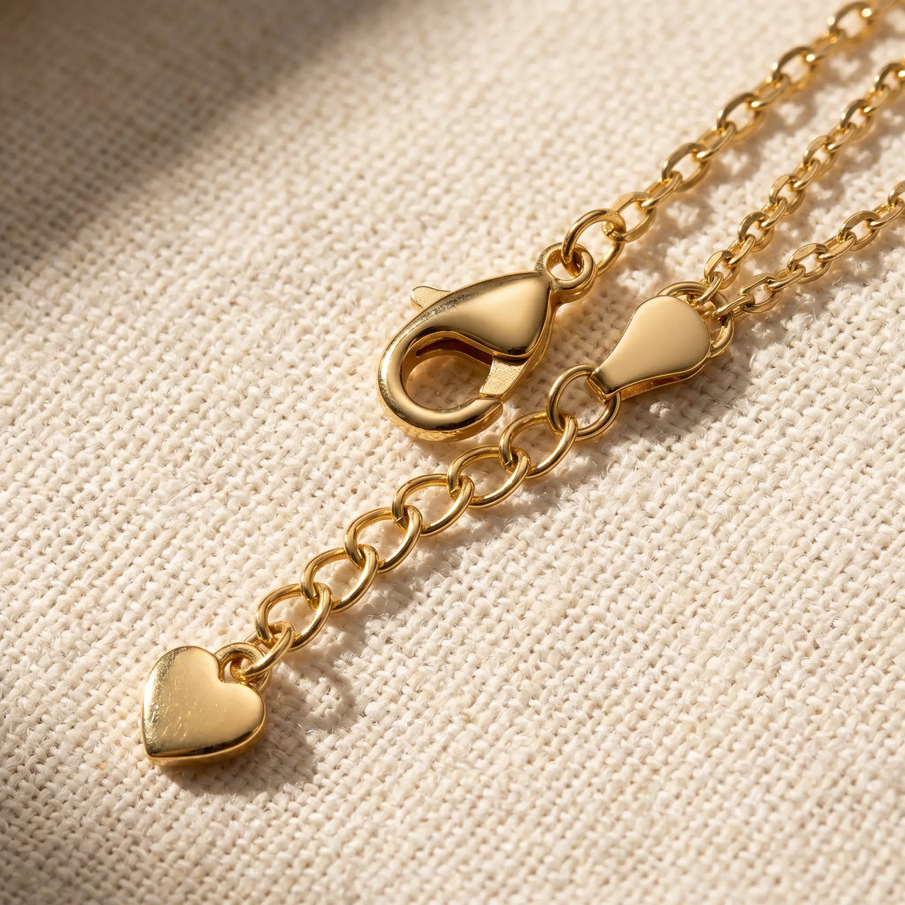 Amalfi Layered Anklet macro detail — polished gold lobster clasp + extension chain + heart-charm pendant tag on cream linen, B&S signature hardware
