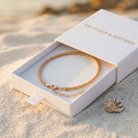 Thumbnail for Maëlle gold tube-mesh anklet inside B&S signature white slide-drawer gift box on beach sand at golden hour with sea shell — luxury anklet packaging