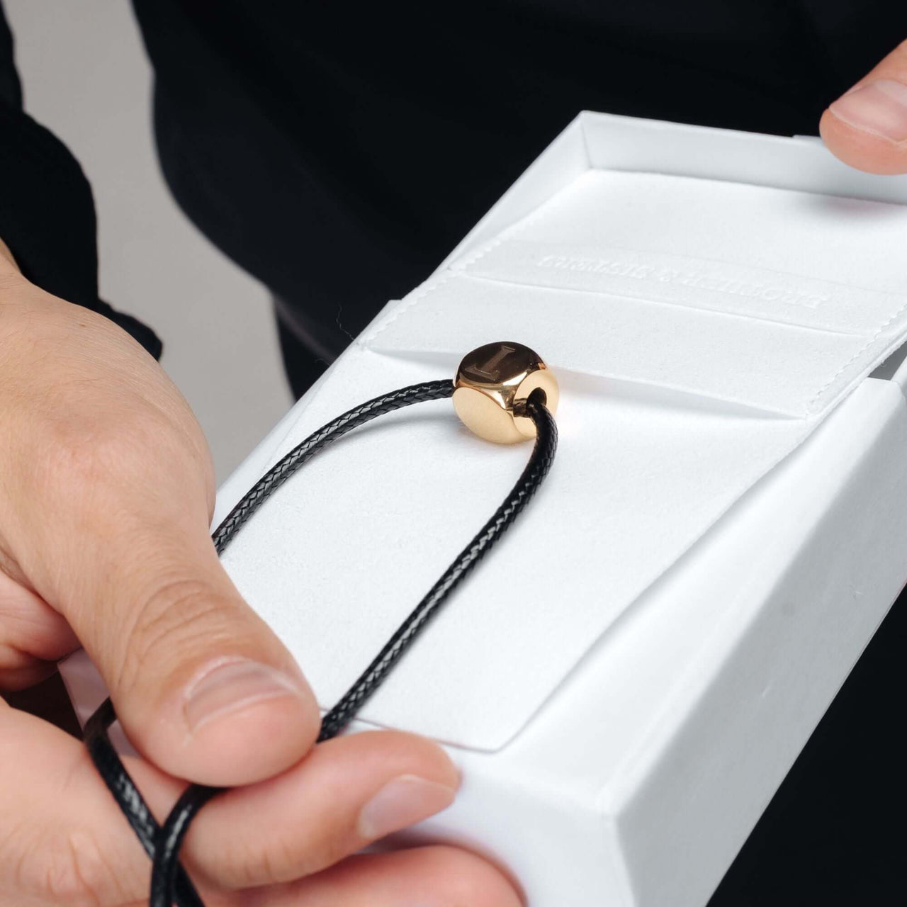 Cube Initial Necklace held in hand, featuring a gold cube pendant and black cord, by Brother & Sisters.